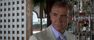 A middle-aged James Bond is standing, half-smiling, in front of a display of bottles and glasses. He is clearly at a bar outdoors.