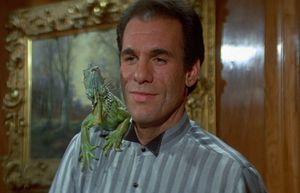 A large dark-haired man stands in front of an ornately framed picture on a panelled wall. He is wearing a nice suit and has an iguana sitting on his shoulder.