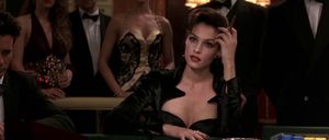 A beautiful white woman with dark red lipstick and a low-cut black dress is sitting at a table in a casino with a cigar in her hand.