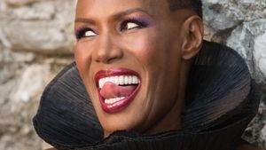 A close up of a black woman in strikingly coloured makeup with a high black collar; she has short hair and her tongue is touching her top front teeth. It's May Day.