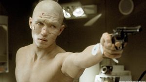 A beautiful white-skinned man is standing in a room with instruments in it. He is hairless and shirtless, with a well-defined body and cuts on his face. He is pointing a gun at someone.