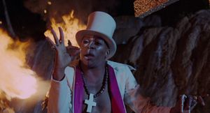 Standing in front of a bonfire is an African man, with half his face painted white. He is shirtless, but wearing a white top hat and a white jacket.
