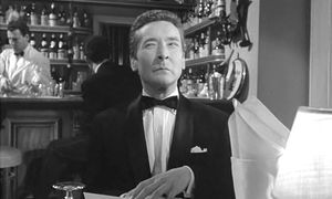 A black-and-white photo of Kenneth Williams in a tuxedo sitting in  a bar. His eyes are narrowed and he looks particularly annoyed.