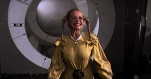 A short young woman with blond plaits wearing glasses and a yellow jumpsuit; she is smiling and standing in front of the door on a space station.